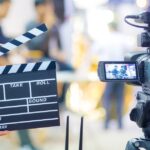 A person holding a clapperboard in front of a recording video camera as it captures two people sitting in chairs.
