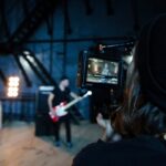 A shot of the back of a person's head behind a camera, recording a drummer and a man playing guitar in the background.