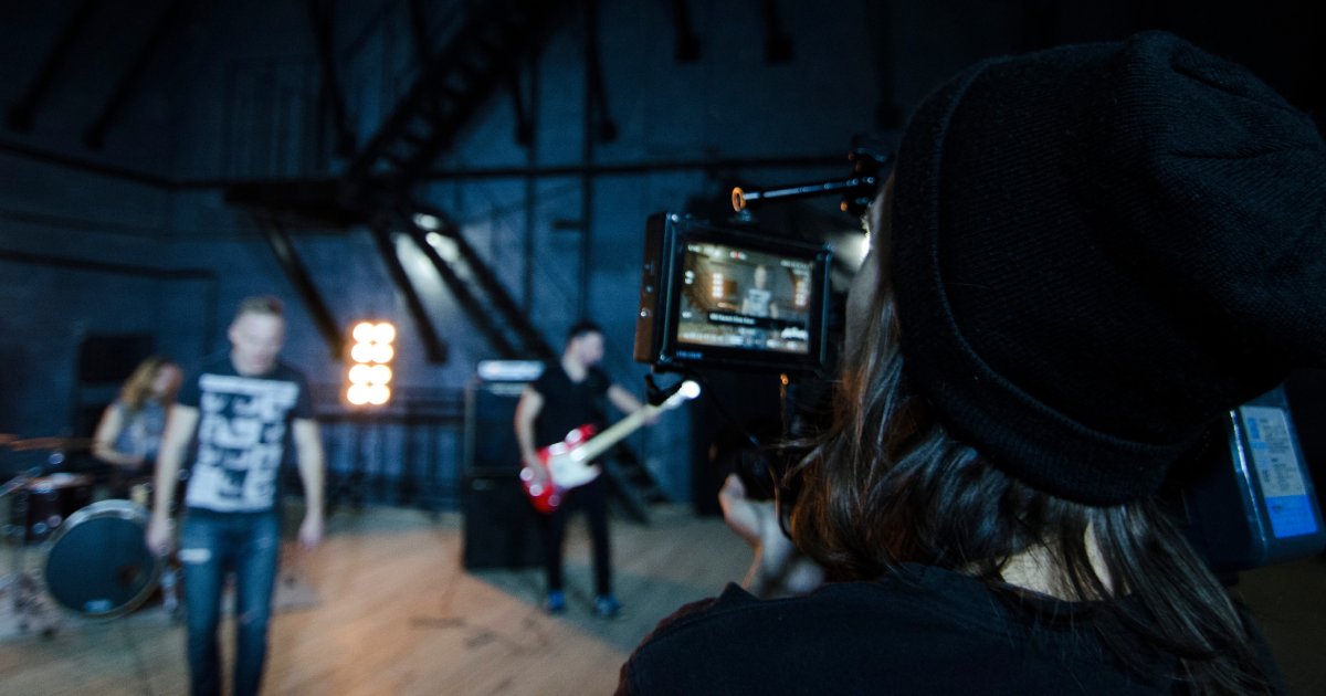 A shot of the back of a person's head behind a camera, recording a drummer and a man playing guitar in the background.