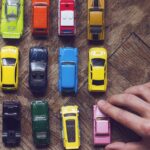 A person places a pink mini model car in their collection. The assorted colored collection sits on a wooden table.