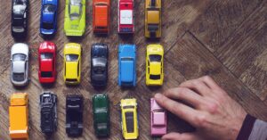 A person places a pink mini model car in their collection. The assorted colored collection sits on a wooden table.