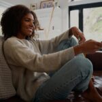 A Black woman sitting on a low couch with her legs crossed. She's smiling and pointing a remote forward.