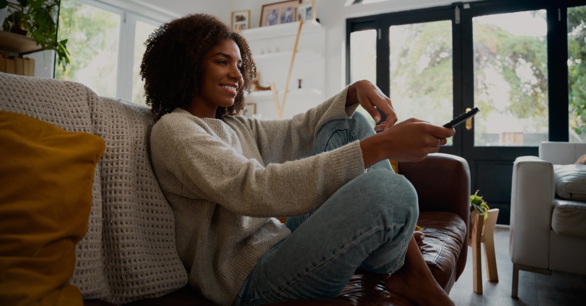 A Black woman sitting on a low couch with her legs crossed. She's smiling and pointing a remote forward.