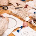 A person sits at a work table with their needle stitch supplies and a project in progress. They wear a cable-knit sweater.