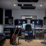 A recording studio with a black rolling chair, two stools, a black couch, two electric guitars, and a large mixing console.