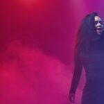 A Black woman stands by a microphone, surrounded by purple and pink lighting with fog in the background.