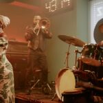 A Black woman in a green dress stands by a microphone, while two drummers and a trombone player perform on stage.