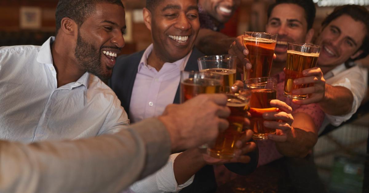 A group of diverse young men smiling and laughing together as they toast pint glasses full of beer in a bar.