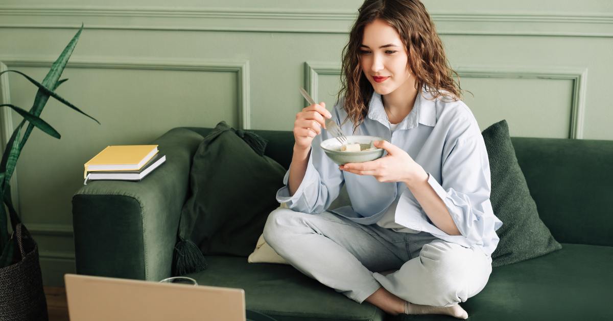 A woman sitting cross-legged on a green couch as she eats from a bowl and watches a laptop across from her.