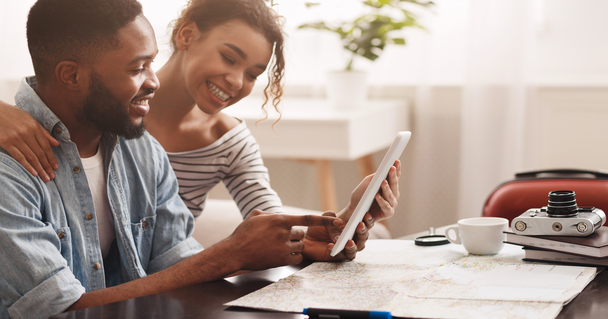A young man and woman smile as they look at an electronic tablet with a road map and old camera in front of them.