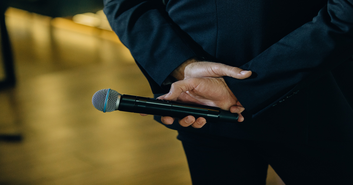 A close-up of the hands of a man wearing a black suit, holding a wireless microphone, standing on a wooden stage.