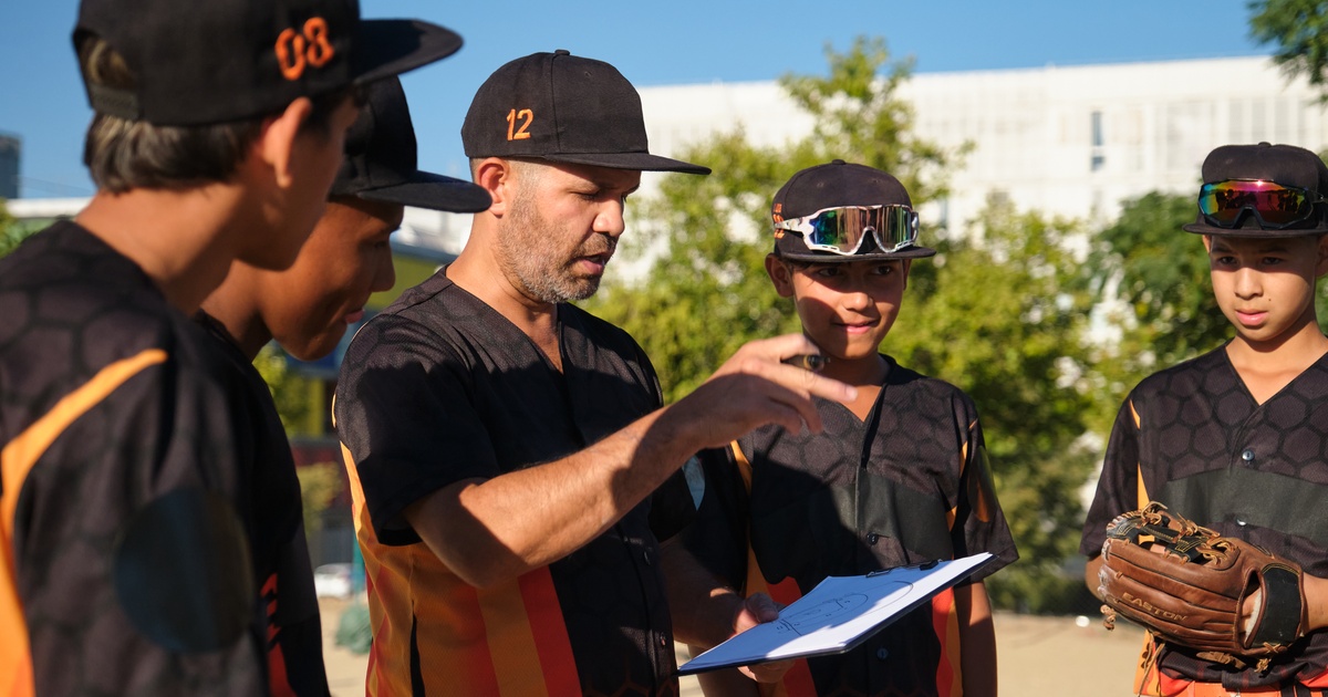 A youth baseball manager in a huddle with his team giving instructions to players while looking at a clipboard.