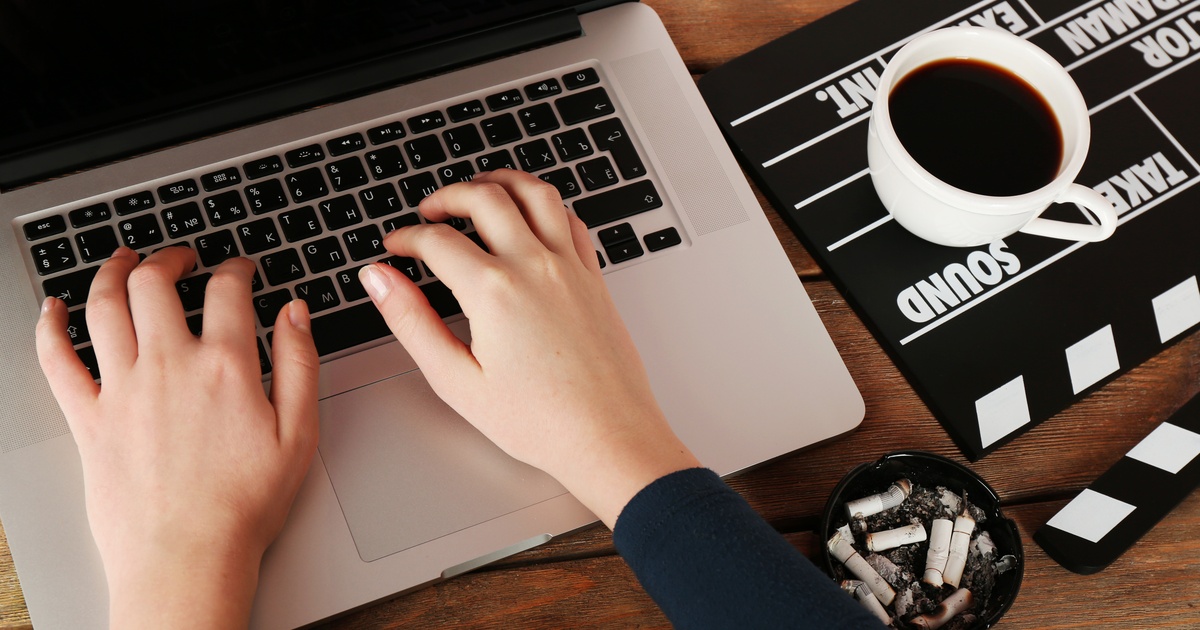 A close-up of someone sitting at their laptop with their hands on the keyboard. A cup of coffee sits on a clapperboard.