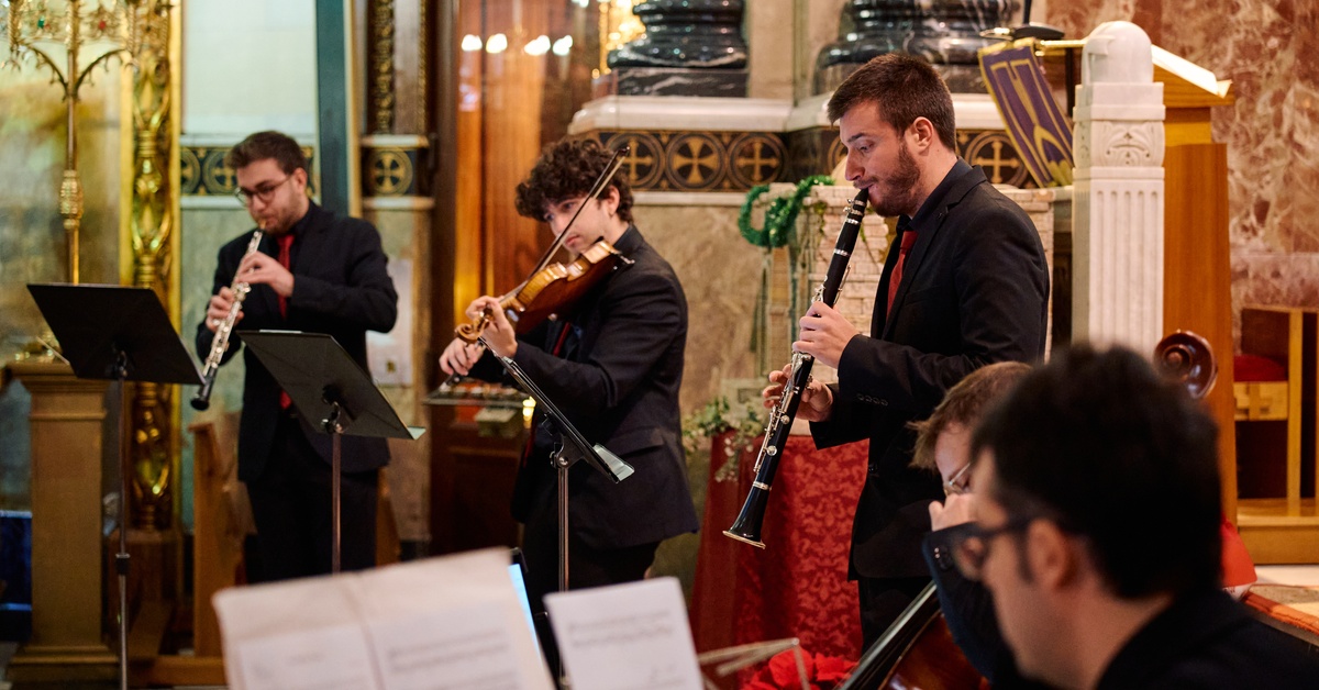 Five musicians wearing black shirts, playing different instruments while looking at partitures inside a church.