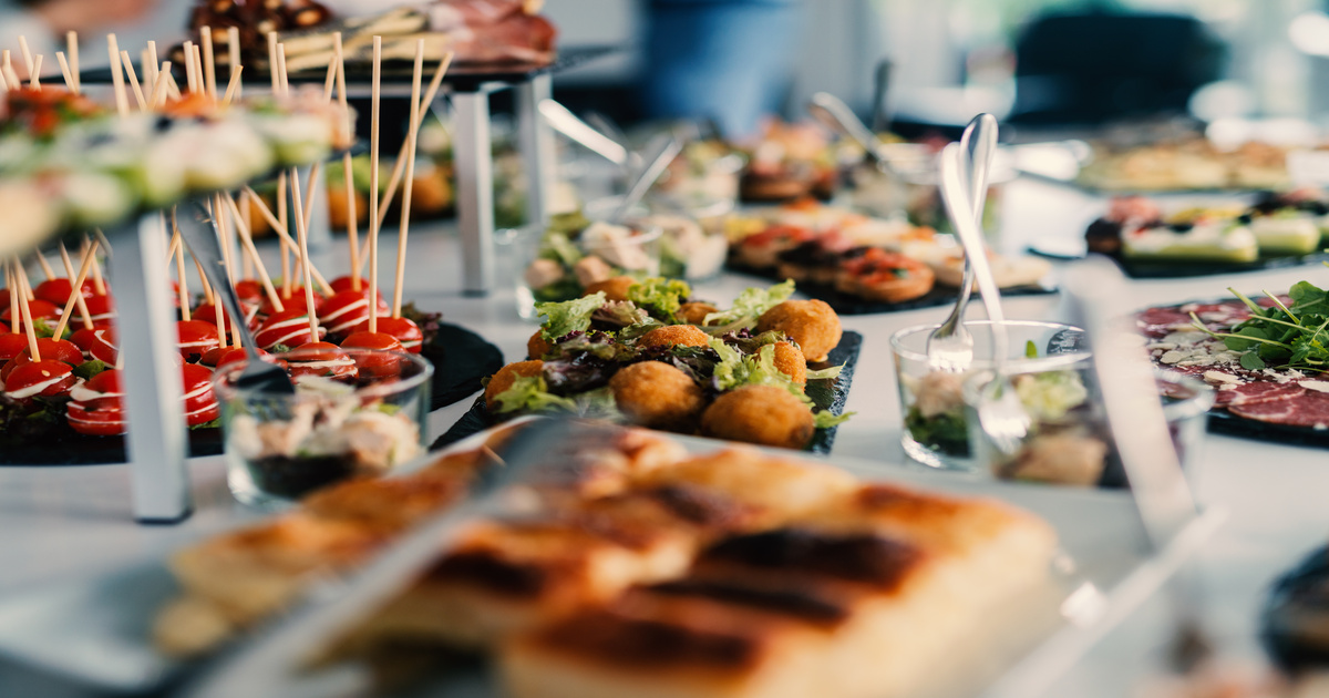 A food station at a large event with various height displays and finger foods spread out. The tablecloth is white.